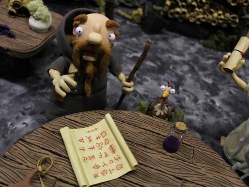 still from stop-motion animation, featuring a druid in a brown robe and a chicken standing at a round wooden table and looking upwards. There is a scroll on the table.