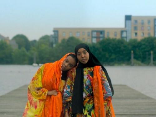 two people wearing orange and yellow diracs, standing on a jetty with a river, trees and buildings in the background.
