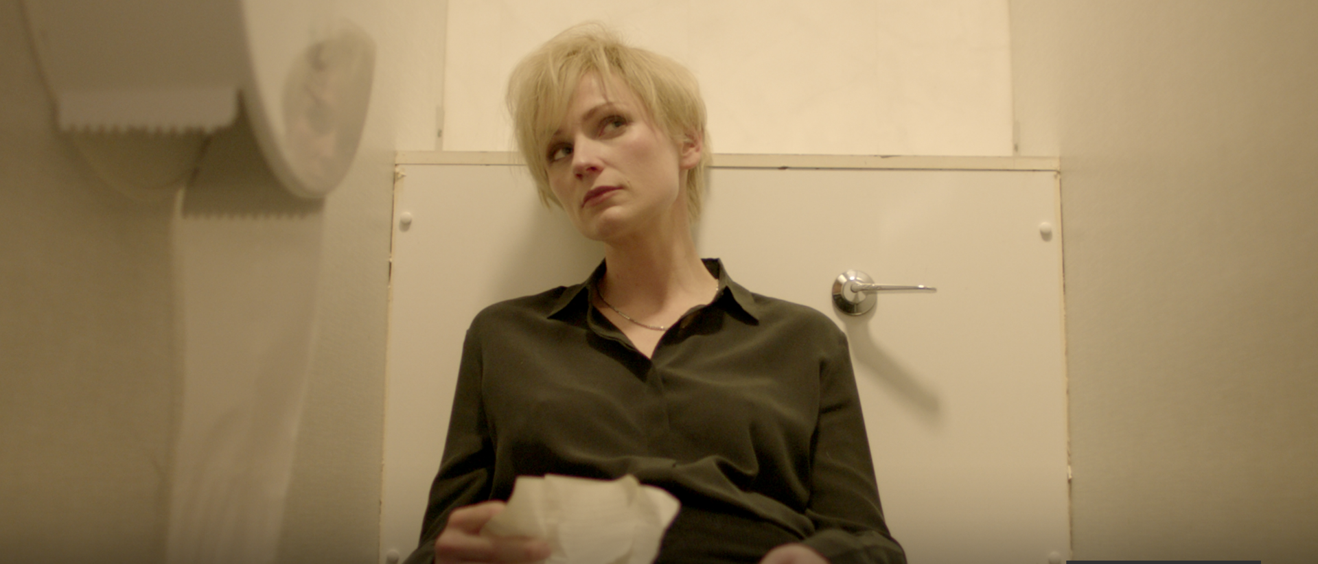 a still from burial featuring a person sitting in a tolet cubicle holding some papers