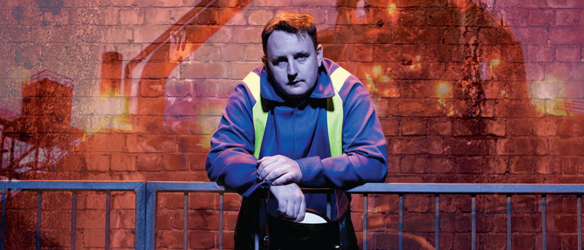 production still from we're still here featuring a man in a steel workers uniform leaning against a metal railing