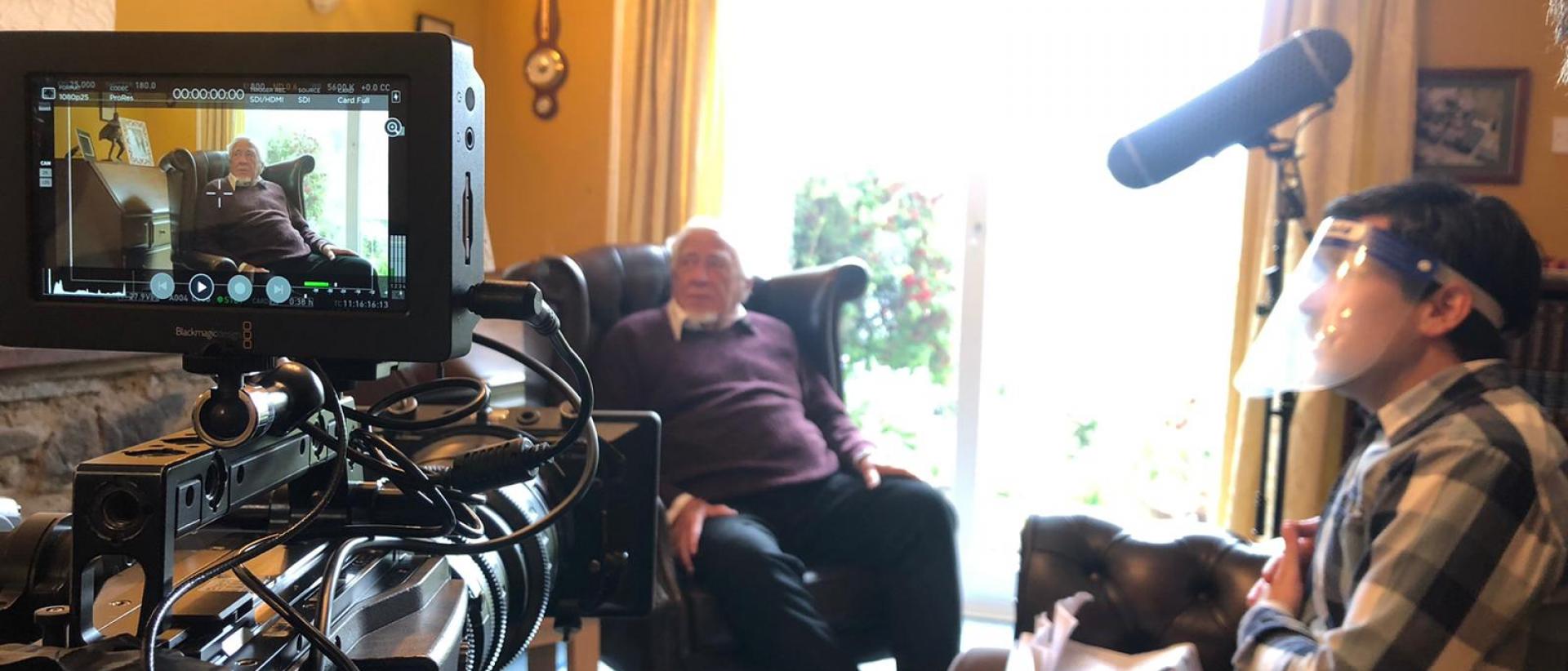 behind the scenes of a short film shoot, featuring a person being interviewed in their living room on camera. The interviewer is wearing a full face covering.