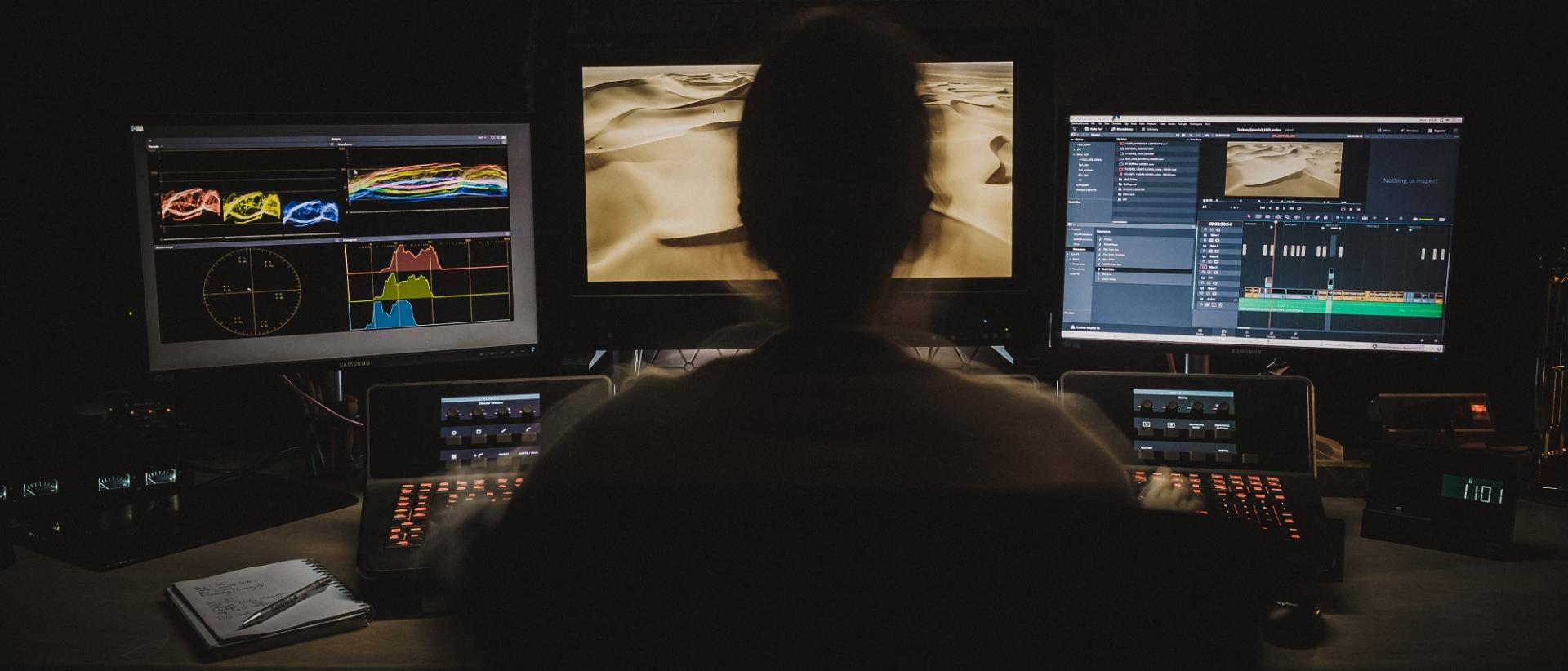 photo of a person sitting in a dark room in front of three computer monitors editing footage.