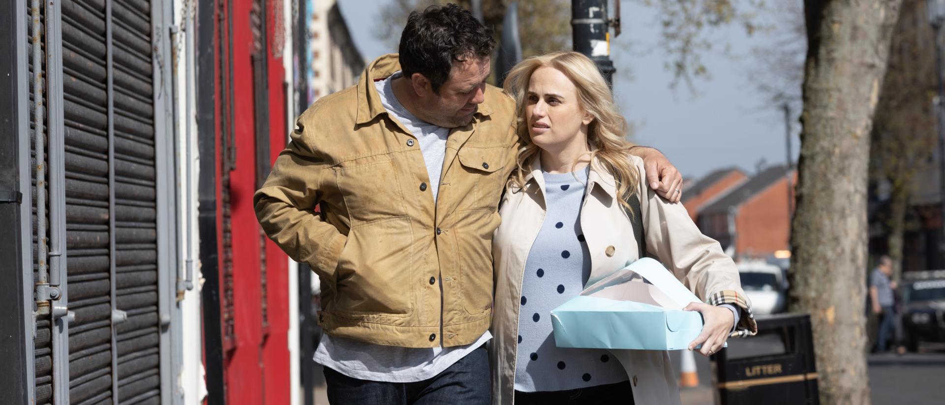still from the almond and the seahorse featuring celyn jones and rebel wilson walking down a street during the day. Celyn has his arm around Rebel's shoulders and she is carrying a box