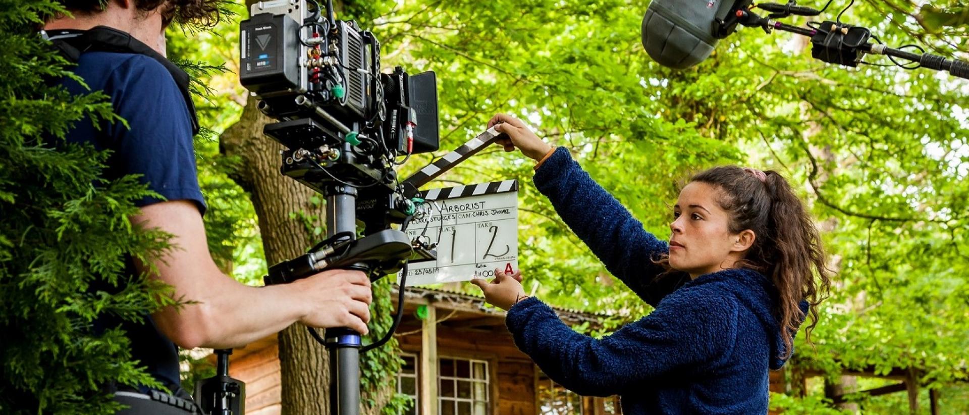 A photo of two people working on a film set outside a wood cabin in a wooded area. 
