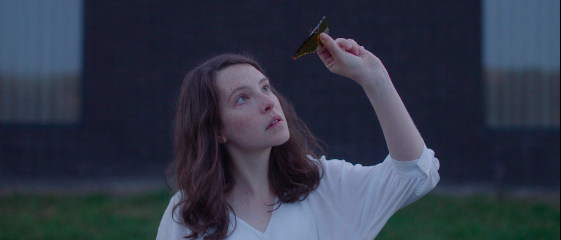 still from Gwledd / The Feast featuring annes elwy wearing a white blouse, standing outside and holding a piece of broken wine bottle up to look at it