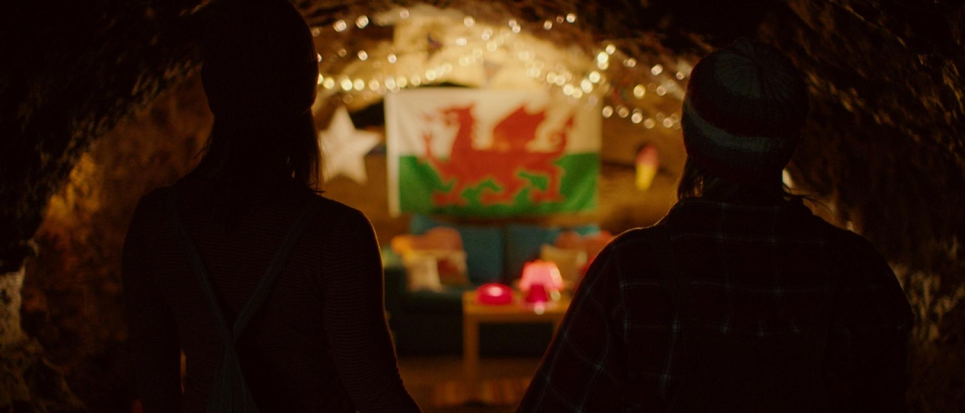 a still from Jelly featuring two people standing in front of a welsh flag in a cave holding hands