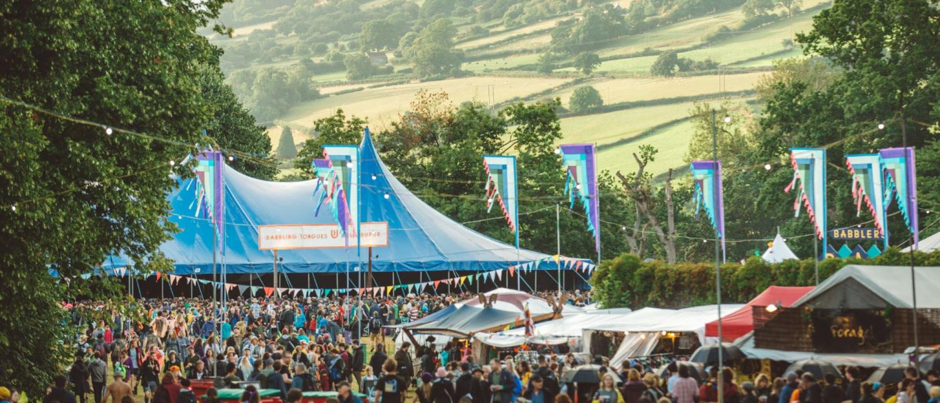 photo from green man festival with lots of people standing in front of a large blue tent. There are trees on a hill in the background.