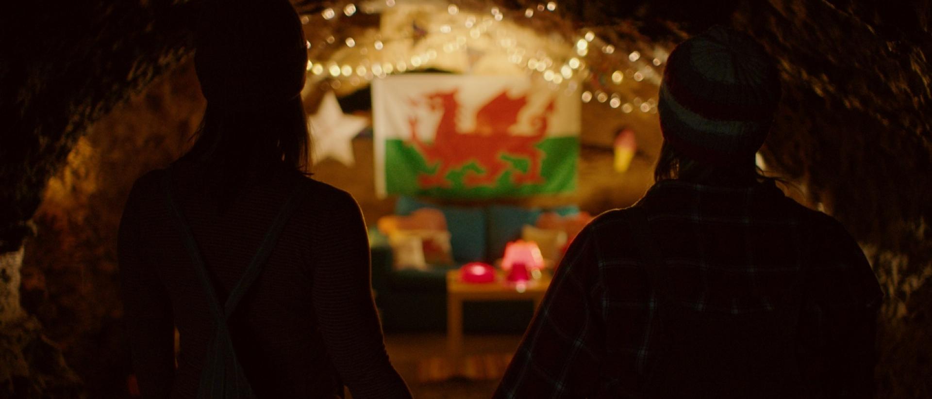 still from jelly featuring two people standing in a cave and holding hands while they look at a welsh flag on the wall, decorated with fairy lights