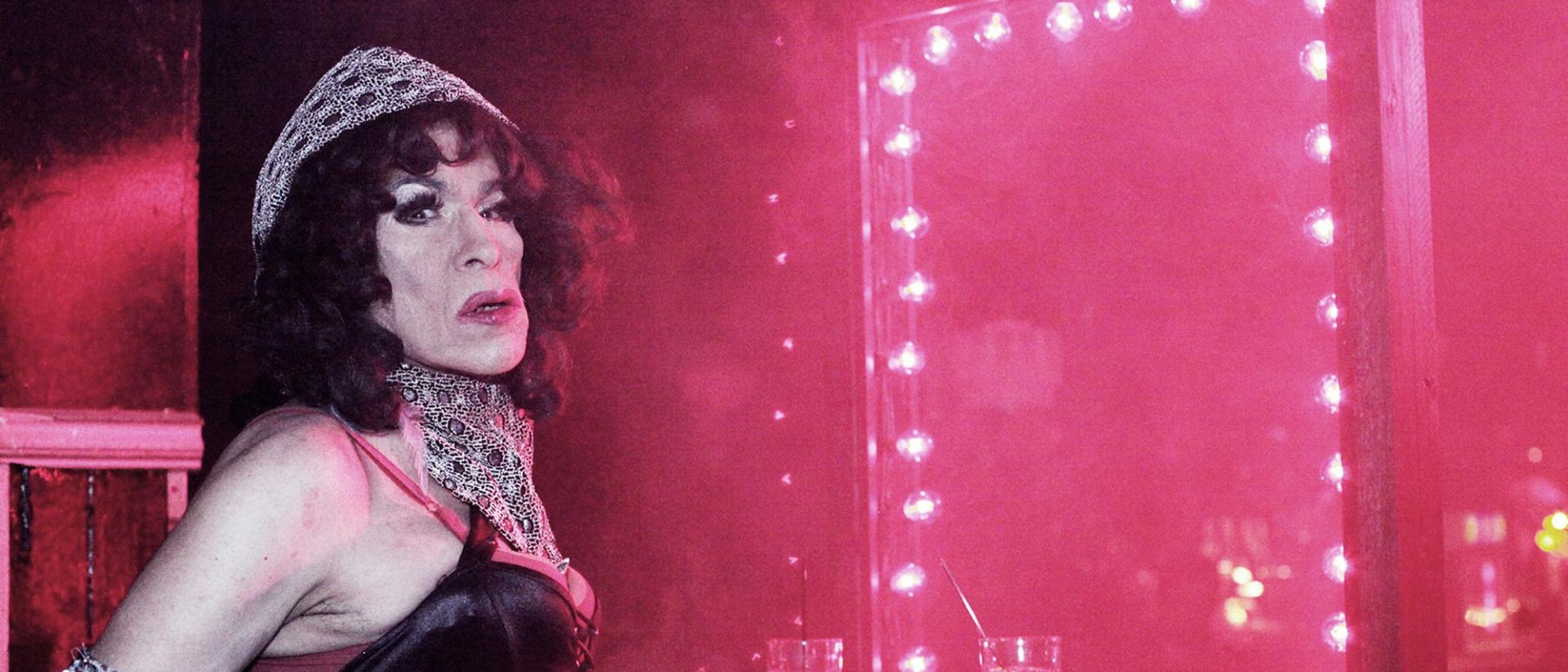 still from donna featuring performer donna personna standing in a dark smoky room next to a mirror with pink lights around the frame