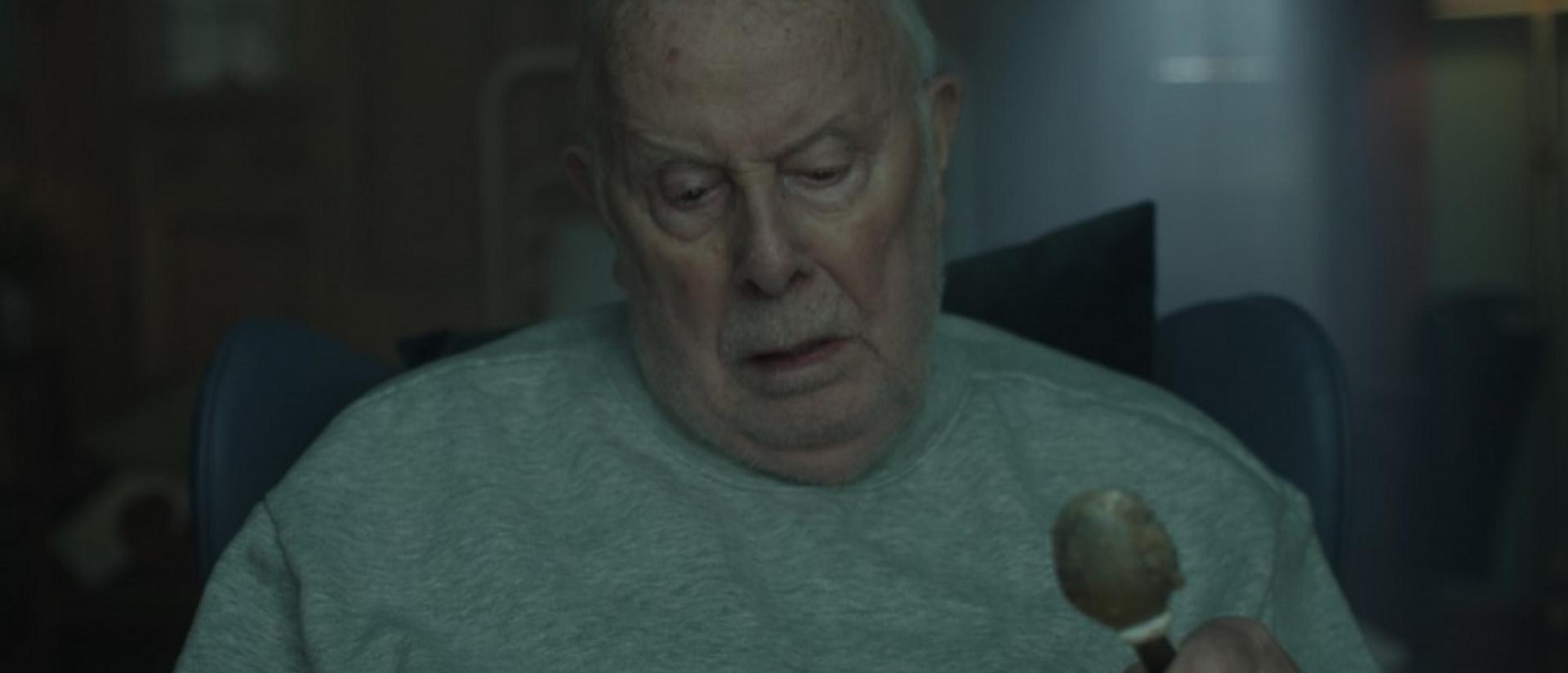 still from g flat featuring an elderly man sitting in a hospital chair, holding a spoon and looking down at a bowl of porridge.