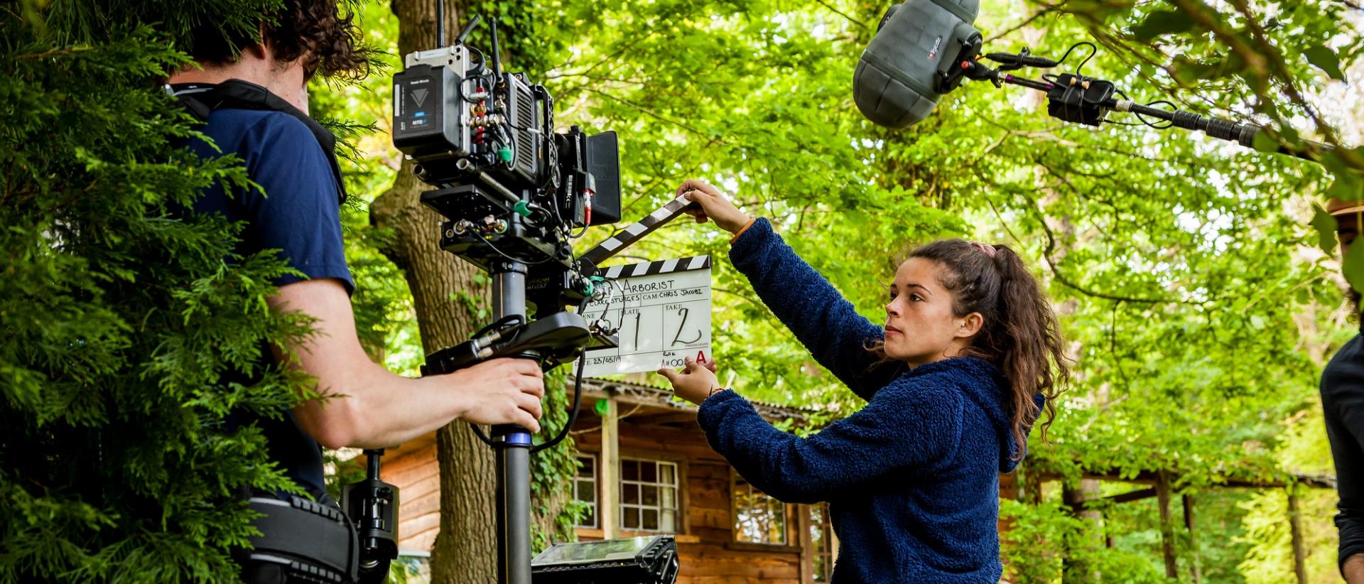 two people working on a film set in a forest. One operates a camera while one holds a clapperboard in front of it.
