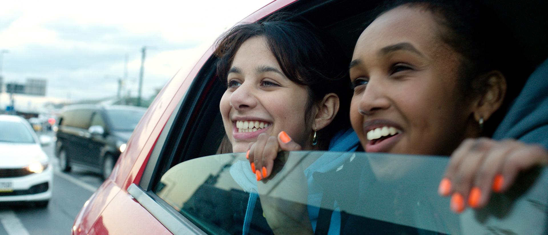 still from brides featuring two teenage girls in the backseat of a car peering out of the half-open window and smiling