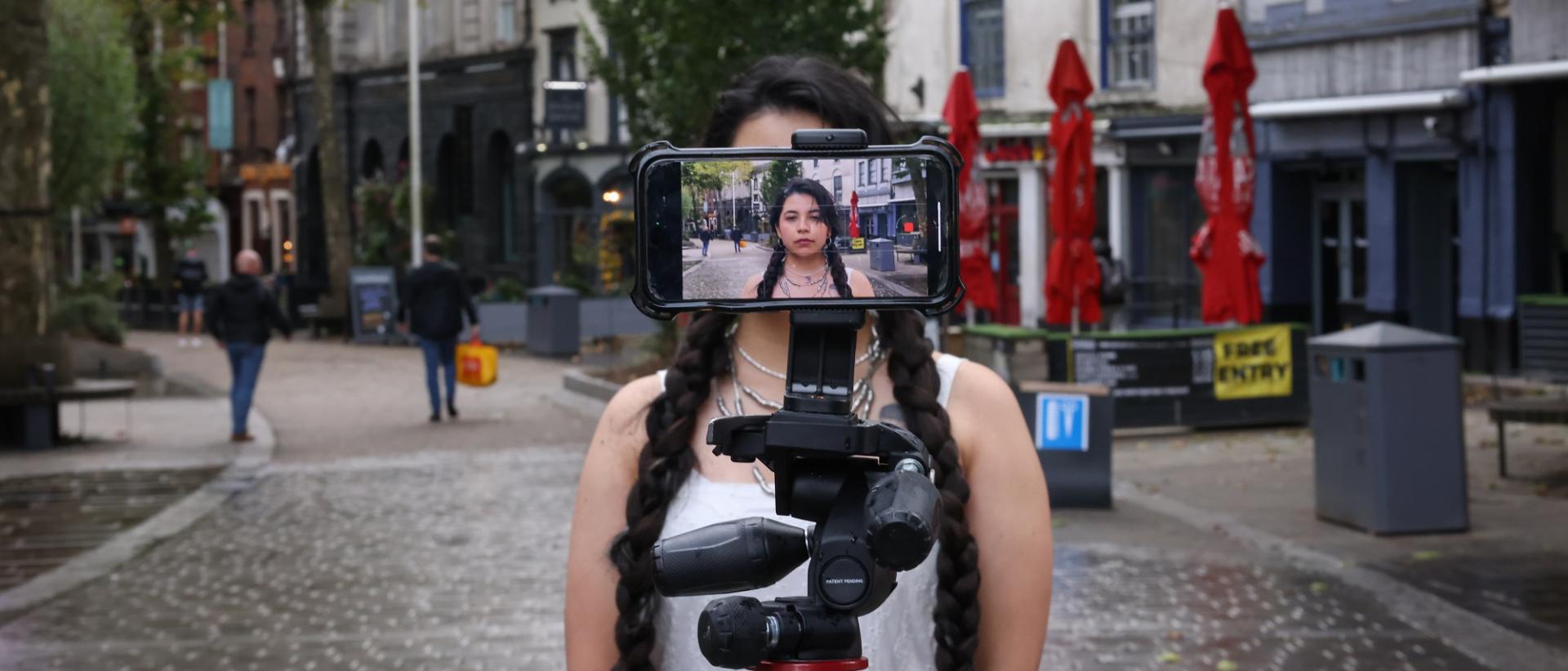 photo of a person standing in a city street in front of a tripod with a smartphone filming them