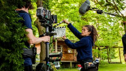 a camera crew shooting in a green forest in front of a wood cabin