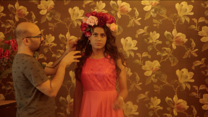 A still from Being Hijra featuring a person wearing a pink dress and flowers in their hair standing in front of a wall with brown floral wallpaper. Another person is standing next to them doing their hair.