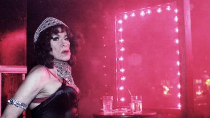 still from donna featuring performer donna personna standing in a dark smoky room next to a mirror with pink lights around the frame