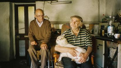 still from forest coal pit featuring two elderly men sitting in a farmhouse kitchen. One has a sheep on his lap.