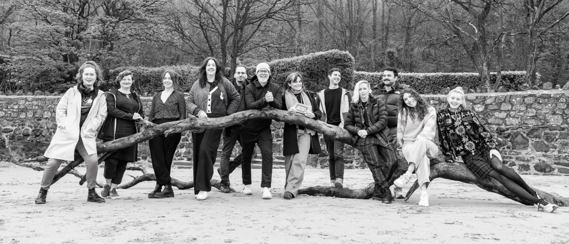 black and white photo of 12 people posing on a beach by a fallen tree trunk. behind them is a low stone wall and trees